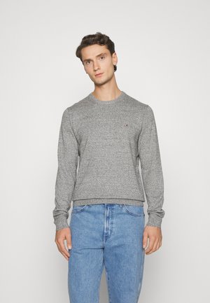 Maglione grigio lavorato a maglia con scollo a girocollo, polsini e orlo a costine. Presenta un piccolo logo sul petto. Abbinato a jeans azzurri chiari.