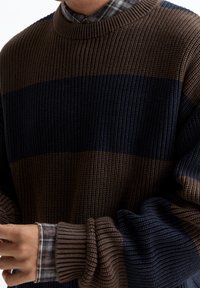Homme portant un pull tricoté à rayures marron et bleu marine par-dessus une chemise à carreaux, ajustant la manche du pull avec ses mains.