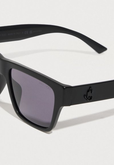 Μαύρα γυαλιά ηλίου με τετράγωνα πλαίσια και γκρι φακούς, με υφή ματ και μικρό μαύρο λογότυπο στο πλάι. Ανθεκτικός σχεδιασμός.