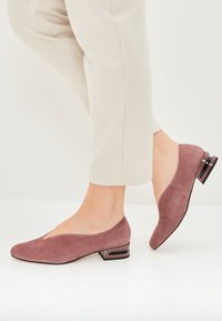Pantofi cu toc conic, din suede, de culoare roz prăfuit, cu vârf ascuțit și părți laterale decupate. Accesoriul din călcâi dreptunghiular negru adaugă un contrast și o estetică modernă.