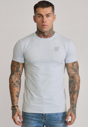Mann med omfattende tatoveringer på armer og nakke, iført en lyseblå tettsittende T-skjorte og blå jeans, vendt mot kamera med ensfarget bakgrunn.