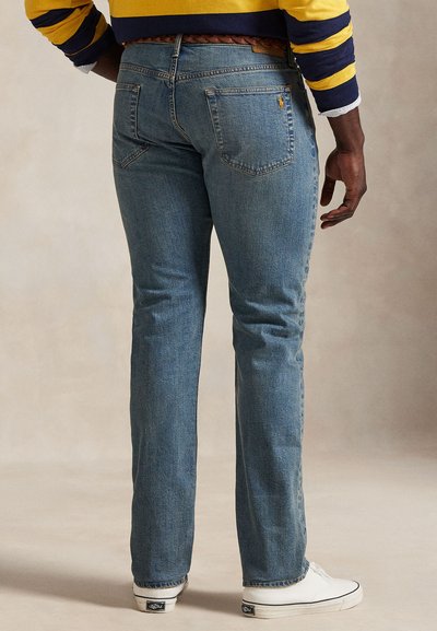 Mand iført lyseblå jeans, hvide sneakers, brunt flettet bælte og en langærmet trøje med gule og marineblå striber, står med ryggen til.