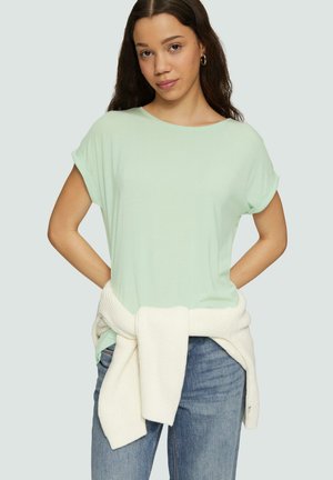 Lichtgroene t-shirt met korte mouwen en een ronde hals, gecombineerd met een witte gebreide trui die om de taille is geknoopt, en denim jeans.