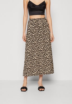 Femme portant un crop top noir, une jupe midi imprimée léopard et des sandales noires tressées à bout ouvert, debout devant un fond uni.