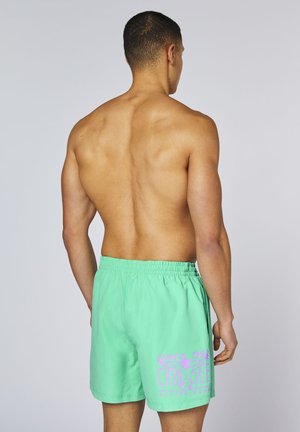 Oberkörperfreier Mann von hinten, trägt hellgrüne Badeshorts mit pinkem Schriftzug und elastischem Bund, steht vor einem schlichten, hellen Hintergrund.