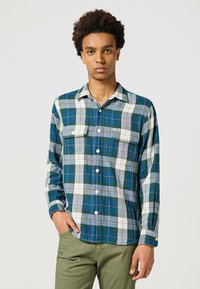 Camisa de cuadros de manga larga en azul, verde y blanco con bolsillos en el pecho. Cuenta con una tapeta de botones y puños ajustables. Hecha de una tela suave.