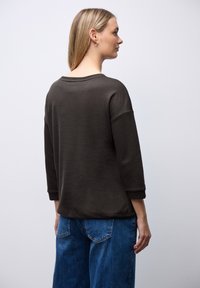 Mørkebrun, langærmet top med en afslappet pasform, der har ribbede detaljer på ærmerne. Kombineret med blå denimbukser. Bagview.