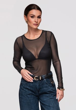 Mujer con cabello oscuro lleva una blusa negra transparente de manga larga sobre un sujetador negro, vaqueros azules, cinturón negro, pendientes de aro dorados y reloj de pulsera sobre fondo liso.