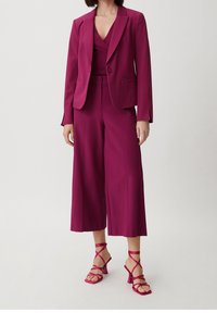 Costume magenta avec un blazer ajusté et un pantalon large, présentant un tissu lisse et un design minimaliste. Associé à des sandales magenta à lanières.