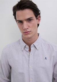 Skjorte med knapper, der har et lys lilla og hvidt stribet mønster samt en lille marineblå kranielogo på venstre brystlomme.