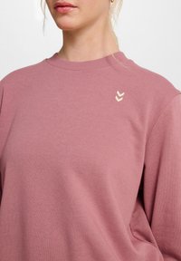 Lyserød sweatshirt i blødt stof med ribbet krave. Har et lille logo i cremefarvet chevron-design på venstre bryst.