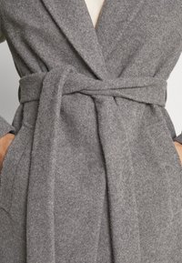 Manteau en laine gris avec une large ceinture nouée autour de la taille, mains dans les poches latérales, et un vêtement intérieur beige visible au niveau de l'encolure.