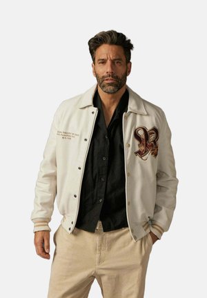 Homme barbu portant une veste blanche brodée, une chemise noire et un pantalon beige, debout avec une main dans la poche.