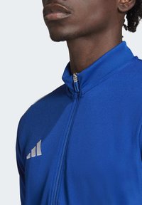 Blue zip-up jacket with a high collar, featuring a smooth texture, silver accents, and logo detail on the chest.