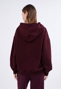 Felpa bordeaux, in misto cotone, vestibilità oversize, grande cappuccio, spalle scese, polsini e orlo a coste, texture morbida, design minimalista.