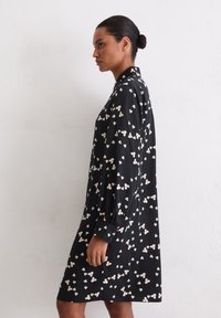 Czarna sukienka w kwiaty z długimi rękawami, zdobiona białym kwiatowym wzorem, z kołnierzykiem i lekko luźnym krojem. Miękka tekstura materiału.