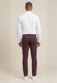 Mand stående med ryggen til, iført en hvid langærmet skjorte, bordeaux bukser med baglommer og hvide sneakers på en beige baggrund.