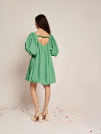 Mulher com cabelo longo e escuro, vestindo um vestido curto verde com mangas bufantes e costas abertas, saltos plataforma bege, de pé sobre confetes espalhados.