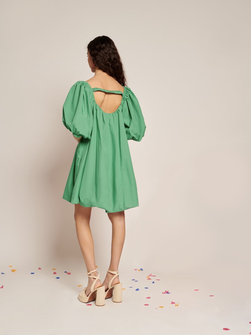 Mulher com cabelo longo e escuro, vestindo um vestido curto verde com mangas bufantes e costas abertas, saltos plataforma bege, de pé sobre confetes espalhados.