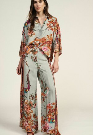 Donna che indossa una blusa larga azzurro chiaro con motivo floreale e paisley e pantaloni a gamba larga con disegni colorati di uccelli e fiori.