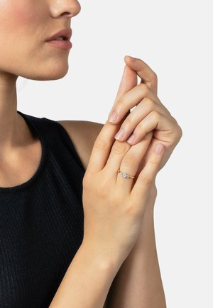 Goldring mit drei funkelnden Edelsteinen. Die Hand des Modells hält sanft die Finger und zeigt die glatte Haut sowie ein dunkles, geripptes Tanktop.