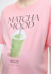 Άτομο που φοράει ροζ πουκάμισο με το κείμενο "MATCHA MOOD" και εικόνα με πράσινο ρόφημα matcha, και το σλόγκαν "ΤΟ ΠΡΑΣΙΝΟ ΕΙΝΑΙ ΤΟ ΝΕΟ ΜΑΥΡΟ".