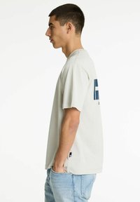 Camiseta de algodón gris claro con manga corta, corte relajado, que presenta un diseño impreso en azul en la parte posterior. Etiqueta lateral cerca del dobladillo.