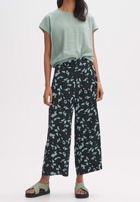 Světle zelený top s krátkým rukávem, volný střih; černé kalhoty s širokými nohavicemi a tyrkysovým květinovým vzorem; zelené sandály s křížovými pásky a silnou podrážkou.