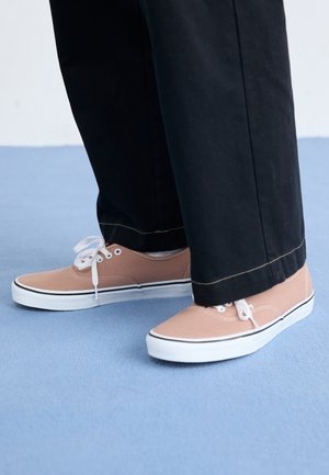 Pieds portant des baskets en toile beige avec des lacets et semelles blanches, associés à un pantalon noir à jambes larges, debout sur un sol bleu clair.