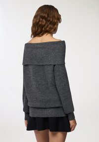 Ciemnoszara, oversize sweter bez rękawów z prążkowanym dekoltem i mankietami, o miękkiej fakturze i luźnym kroju, nałożony na czarną spódnicę.