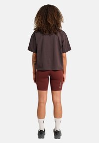Vrouw met krullend haar staat met haar rug naar ons toe, gekleed in een losse donkere shirt, bordeaux kleurige fietsshorts met "ATW" op het linkerbeen, witte sokken en zwarte schoenen.