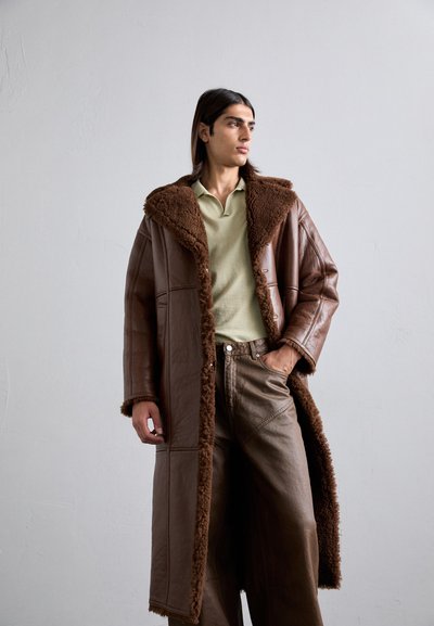 Cappotto di pelle marrone con un collo e una fodera in pelliccia sintetica marrone soffice. Il modello indossa un polo verde chiaro e pantaloni larghi marroni.