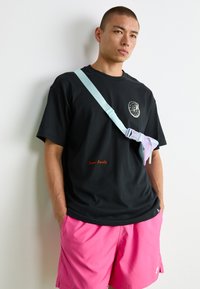 Hombre con camiseta gráfica negra, pantalones cortos rosas y bolso cruzado en tonos pastel, de pie contra una pared blanca con las manos en los bolsillos.
