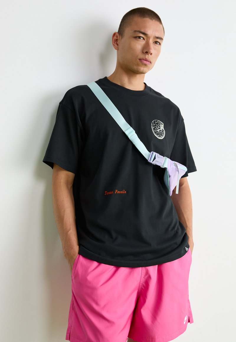 Homme portant un t-shirt graphique noir, un short rose et un sac en bandoulière pastel, se tenant contre un mur blanc avec les mains dans les poches.
