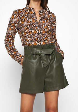 Kobieta ubrana w kolorową, kwiecistą koszulę z długim rękawem, włożoną do wysokich oliwkowozielonych skórzanych szortów z dużym wiązanym paskiem.