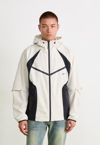 Lettvektet hettejakke i off-white med svarte detaljer, med glidelås, sidelommer og en sporty design med kontrasterende paneler.