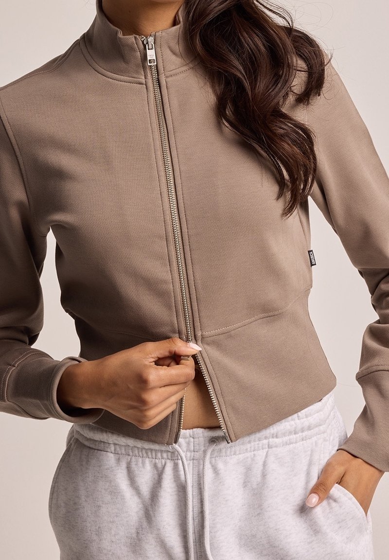 Giacca a zip marrone con colletto alto, design aderente, texture liscia e dettaglio della zip in contrasto, indossata sopra pantaloni della tuta grigi.