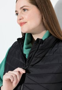 Schwarze gesteppte Steppweste mit hohem Kragen, Reißverschluss und strukturiertem Finish, die über einem grünen Kapuzenpullover getragen wird. Nahaufnahme der Hand am Reißverschluss.