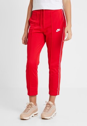 Pantalon de jogging rouge Nike fuselé avec liseré blanc, porté avec des baskets beige Nike sur une personne debout devant un fond uni.