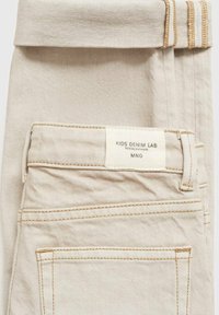 Bež traperice s roliranim pojasom, s oznakom koja glasi "KIDS DENIM LAB" i vidljivim detaljima šivanja.