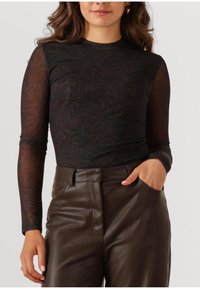 Schwarzes Langarmoberteil mit einem dezenten Blattmuster, aus transparentem Material. Kombiniert mit braunen Lederhosen, die eine glatte Textur aufweisen.