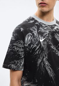 Άνδρας που φοράει μαύρο t-shirt Nike με γκρι αφηρημένα σχέδια που μοιάζουν με φτερά και λευκό λογότυπο Nike στο στήθος, με φωτεινό φόντο.