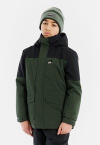 Grüne und schwarze wasserdichte Jacke mit hohem Kragen, seitlichen Taschen, verstellbaren Bündchen und einem Logo auf der Brust. Getragen mit einer passenden Mütze.