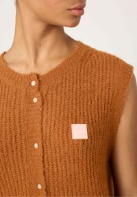 Donkeroranje mouwloze gebreide top met een textuurpatroon, voorzien van decoratieve knopen en een zachte stof. Bevat een lichtroze merketiket.