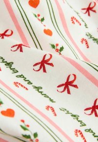 Weißer Stoff mit roten Schleifen, grüner Stechpalme, orangenen Herzen und pinken Zuckerstangen in einem sich wiederholenden Muster; grüne und pinke Streifen akzentuieren das Design.