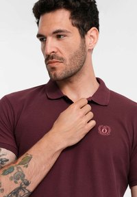 Burgundin värinen t-paita lyhyillä hihoilla, ribbestetty kaulus ja pieni brodeerattu logo rinnassa. Tasainen puuvillasekoitekangas.