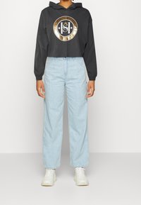 Svart avkortad hoodie med en cirkulär guld- och svart grafisk logotyp, i kombination med ljusblå vida jeans och vita sneakers.