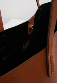 Bruine faux-leder schoudertas met een zwarte binnenkant, voorzien van een drukknopsluiting en inclusief een klein intern merklabel. Gladde textuur.