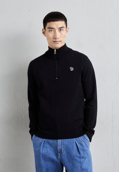 Pull zippé noir avec col montant, présentant un petit design brodé sur la poitrine, associé à un jean en denim bleu clair plissé.