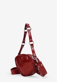 Bolso en forma de corazón de cuero rojo con un acabado brillante, que cuenta con una correa desmontable con herrajes plateados y una textura suave.